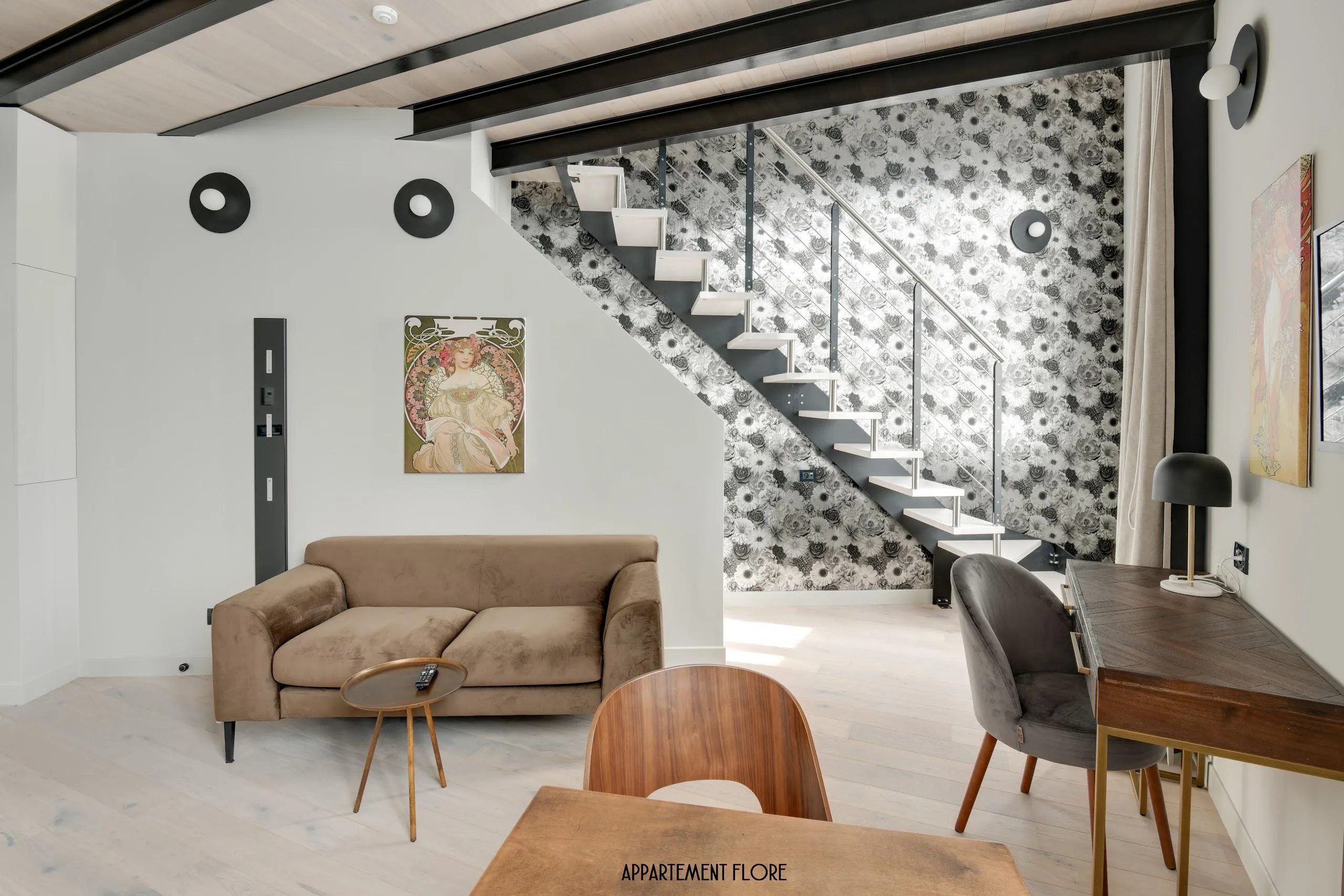 Salon moderne avec canapé marron, table basse, escalier avec papier peint floral noir et blanc.