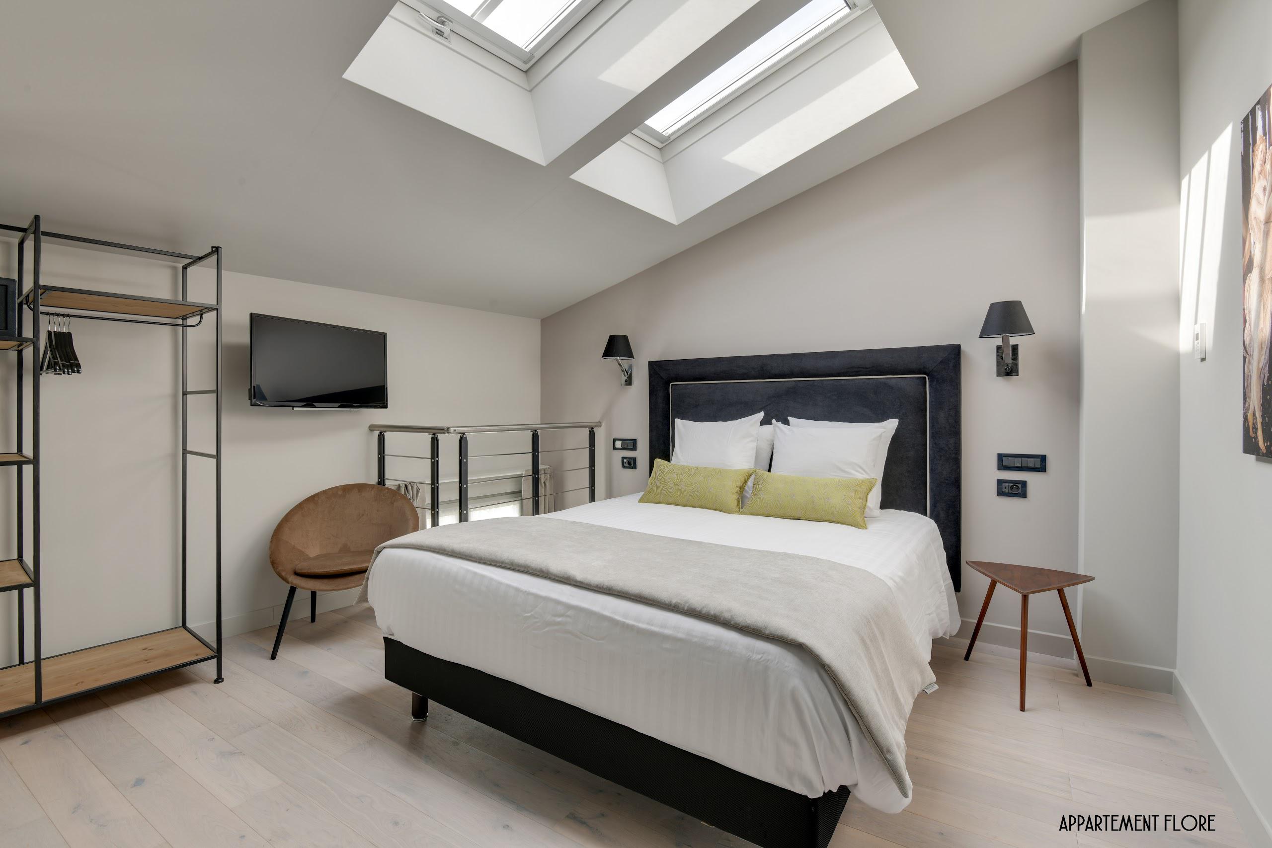 Chambre élégante avec lit double, deux fenêtres de toit, télévision, chaise, et étagères ouvertes.