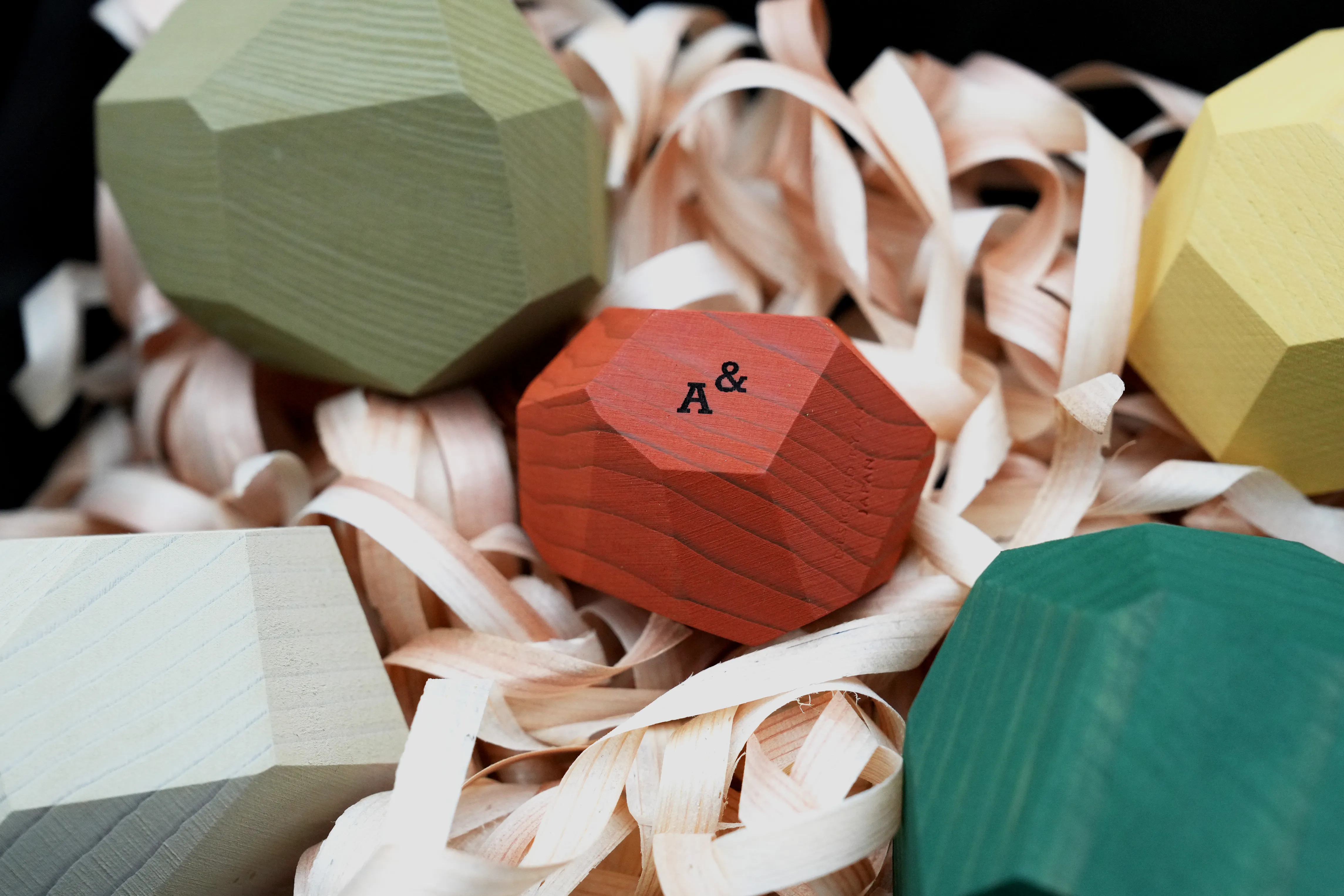 Colorful faceted tumi-isi wooden blocks nested in curled wood shavings, Ampersand branded block in orange