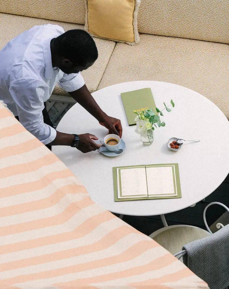 Un homme sert une tasse de café à une table ronde avec un menu, un livre et un vase de fleurs.