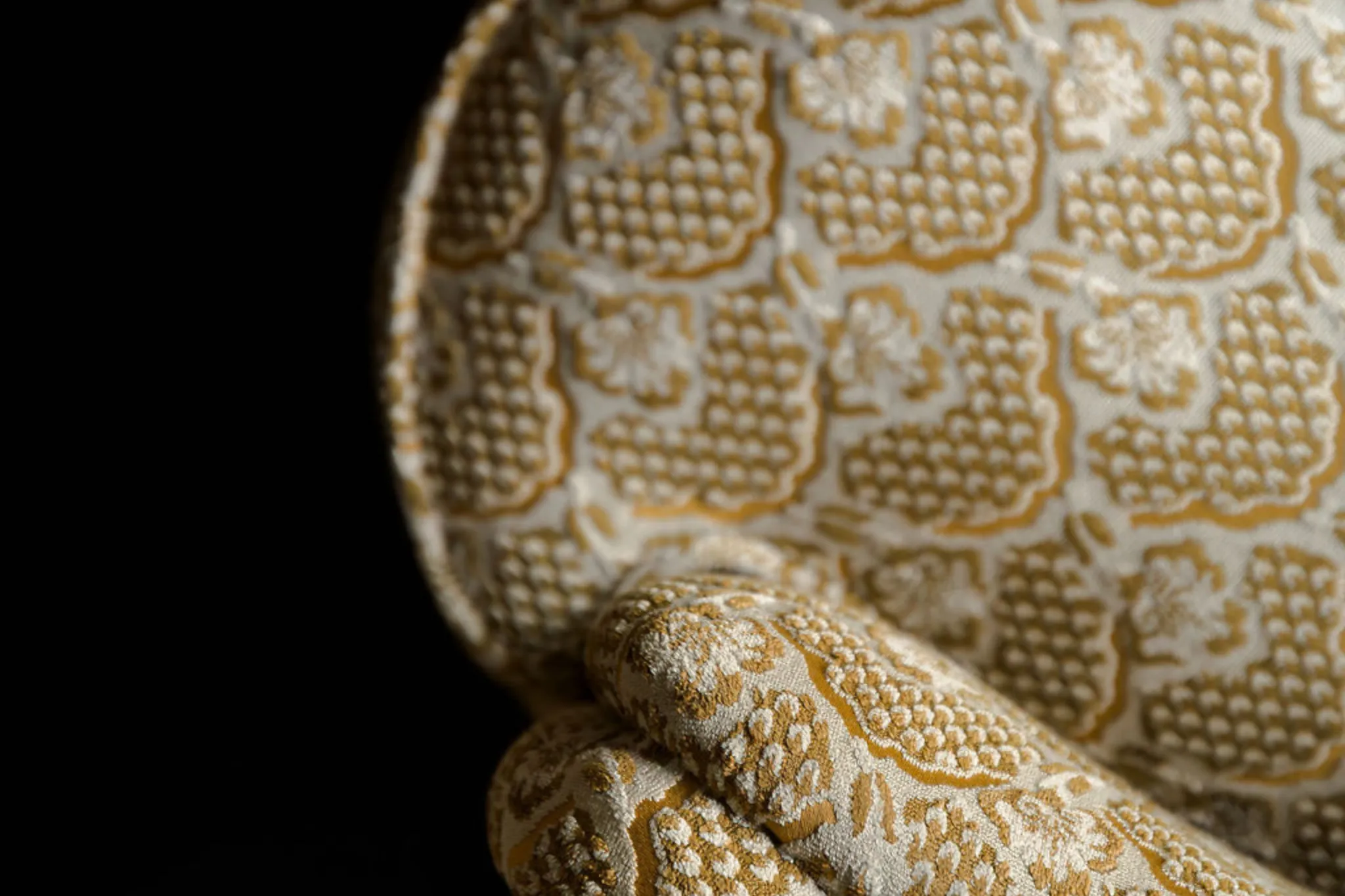 Détail d'un fauteuil avec un tissu beige et motif floral texturé doré sur fond noir.