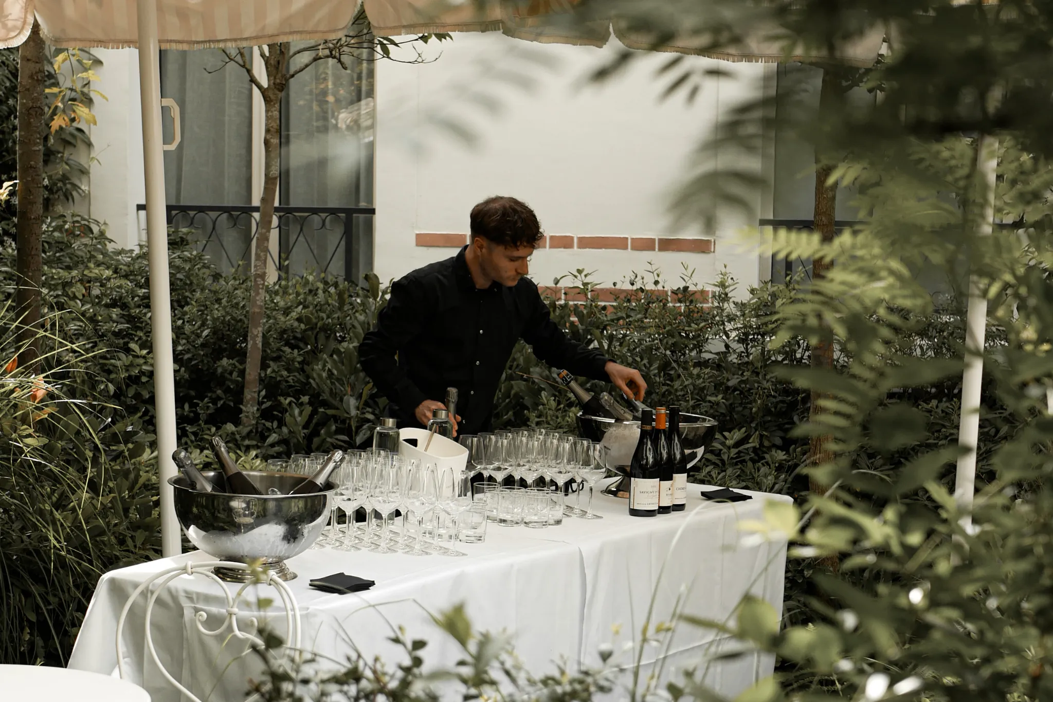 Serveur préparant une table de verre et bouteilles de vin dans un jardin pour événement.