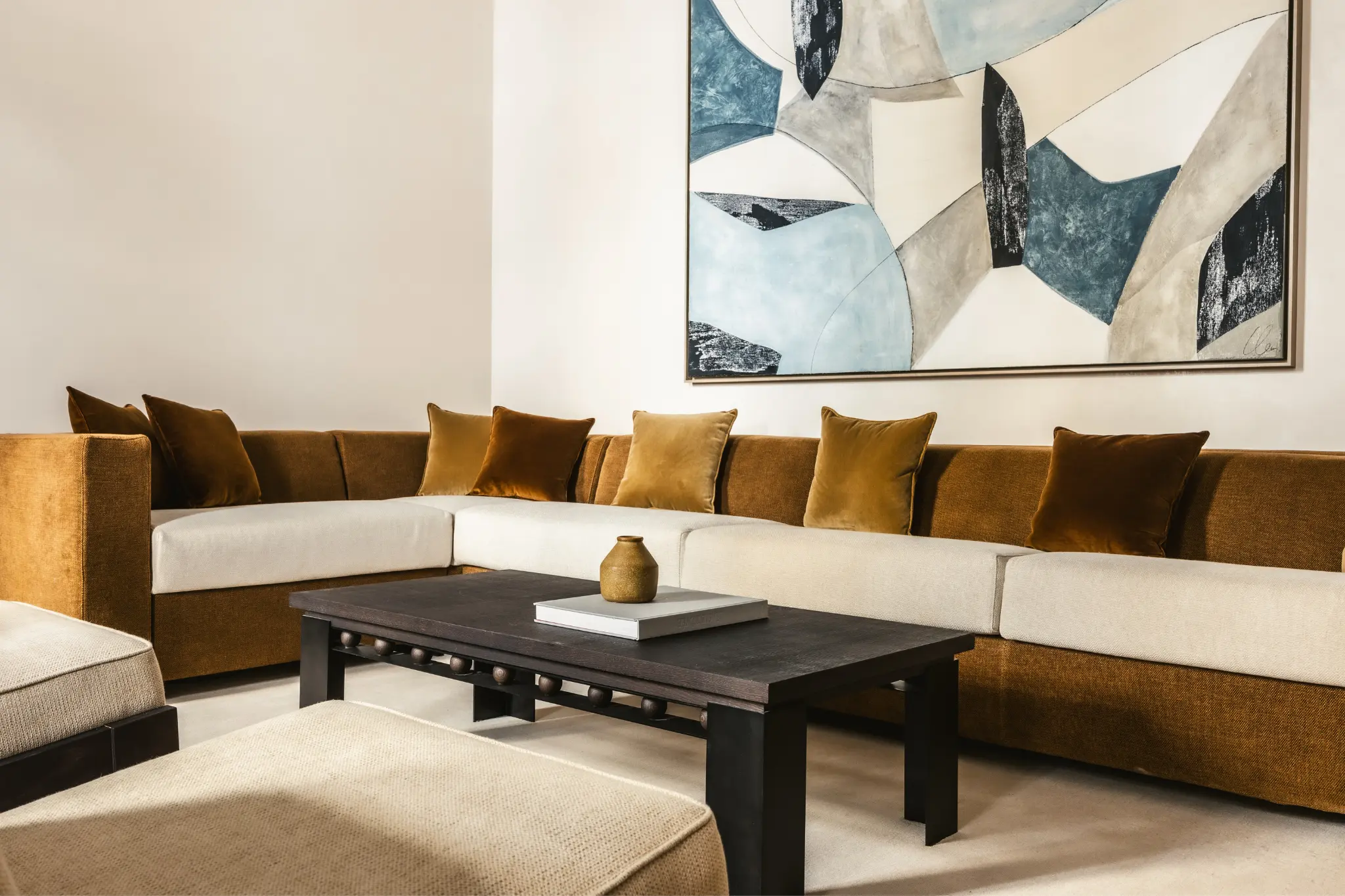 Canapé d'angle marron et beige avec coussins dorés, table basse noire et tableau abstrait bleu et gris.