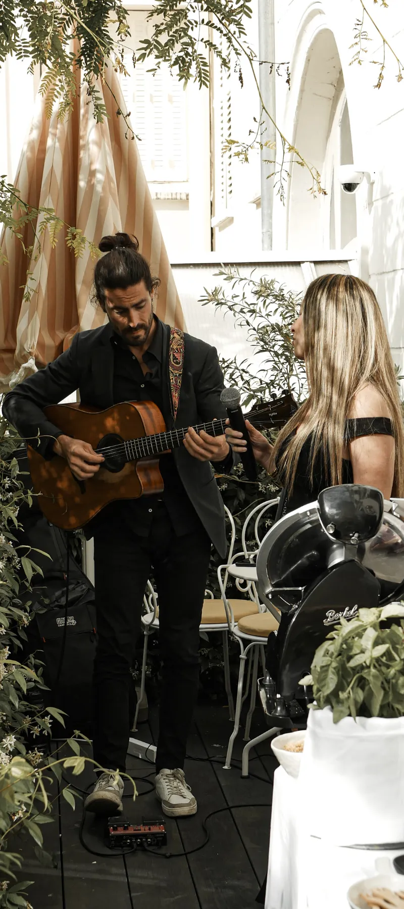 Un homme joue de la guitare tandis qu'une femme tient un micro dans un jardin avec des plantes.