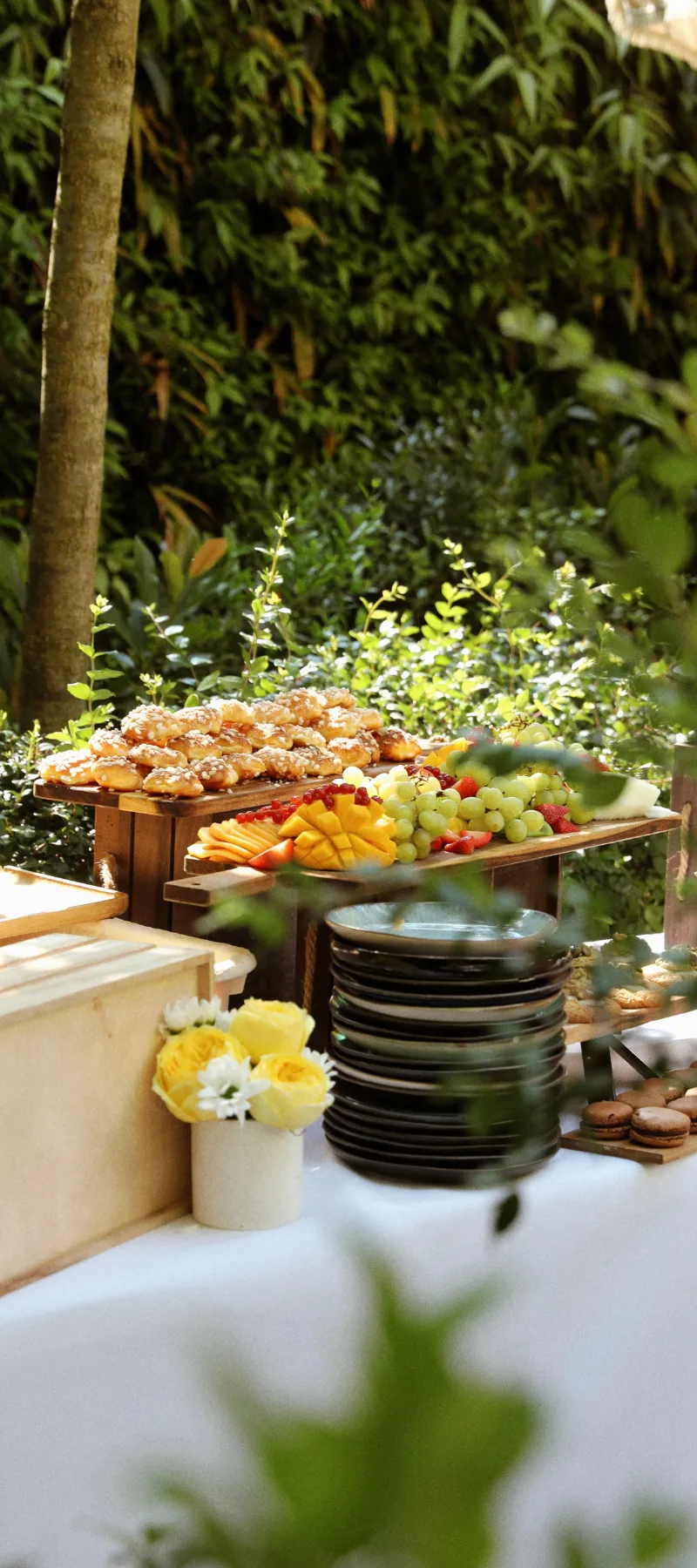 Table en plein air avec plateaux de fruits, pâtisseries, macarons et assiettes empilées.