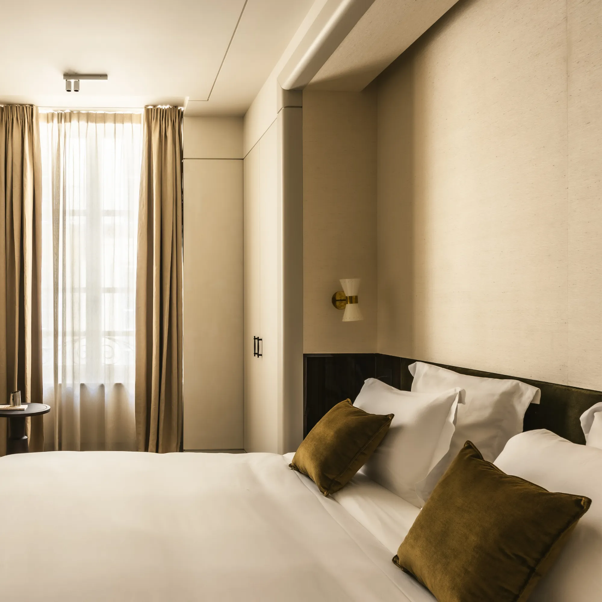Chambre calme avec un lit double blanc, deux coussins marron et rideaux beiges devant une fenêtre.