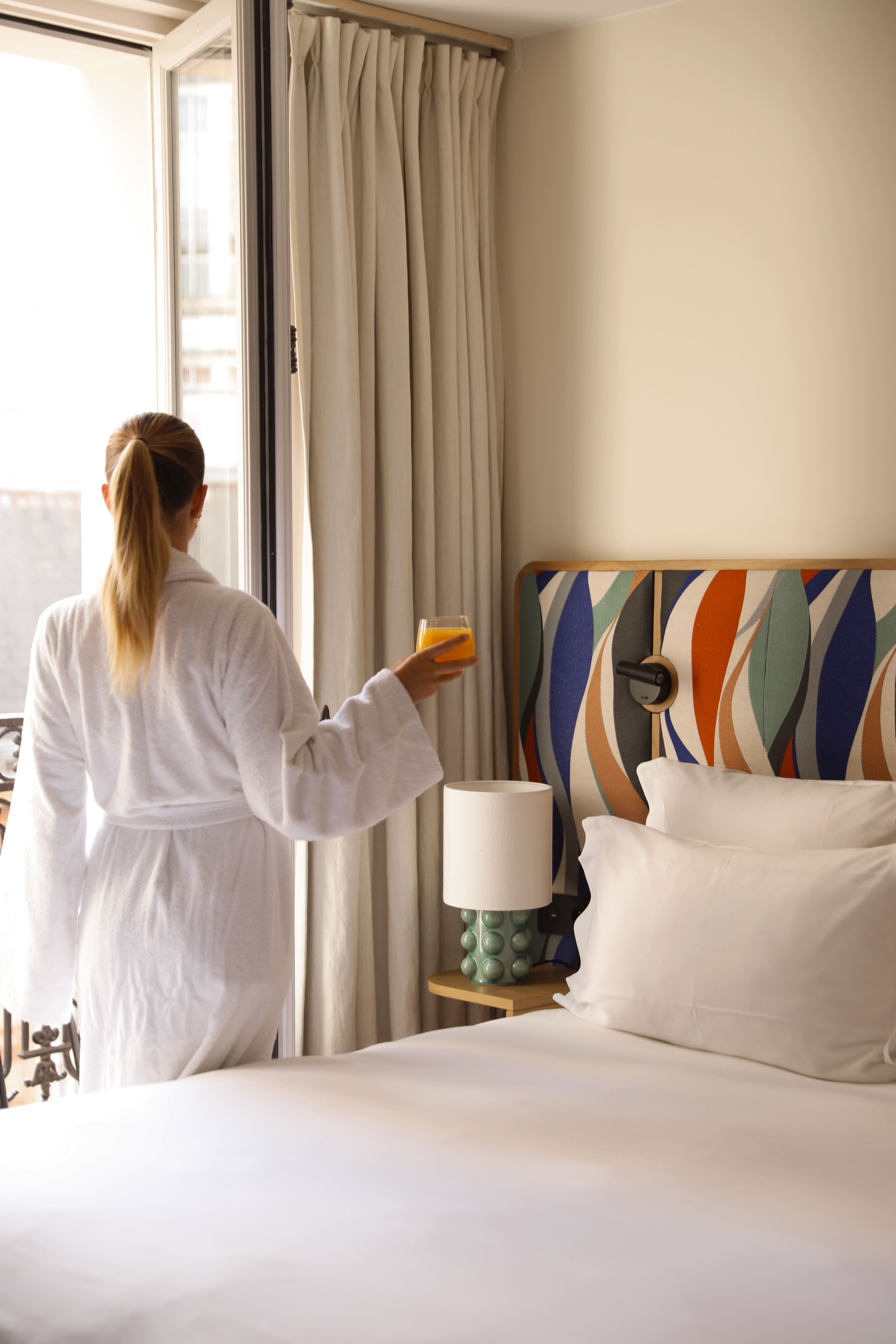 Femme en peignoir blanc tenant un verre de jus d'orange près d'une fenêtre dans une chambre.