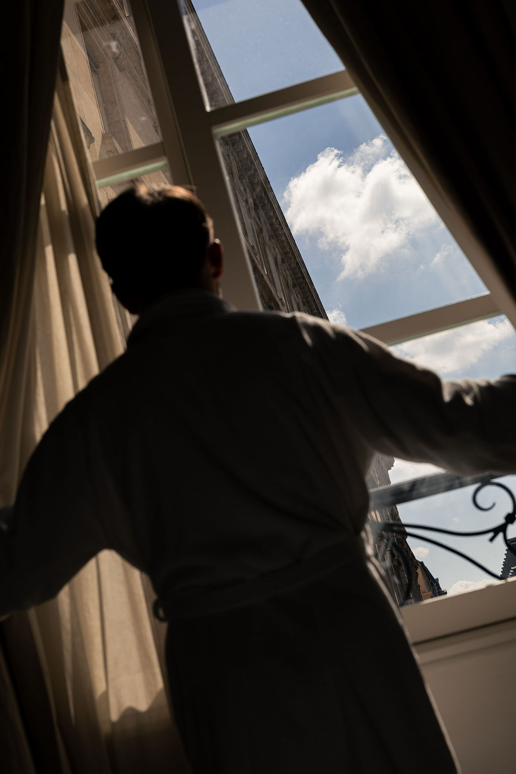 Person vue de dos en robe de chambre regardant par une fenêtre ouverte sur un ciel bleu avec des nuages.