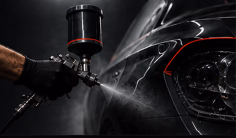 A gloved hand holding a spray gun applying paint to the side of a black car in a dark environment.