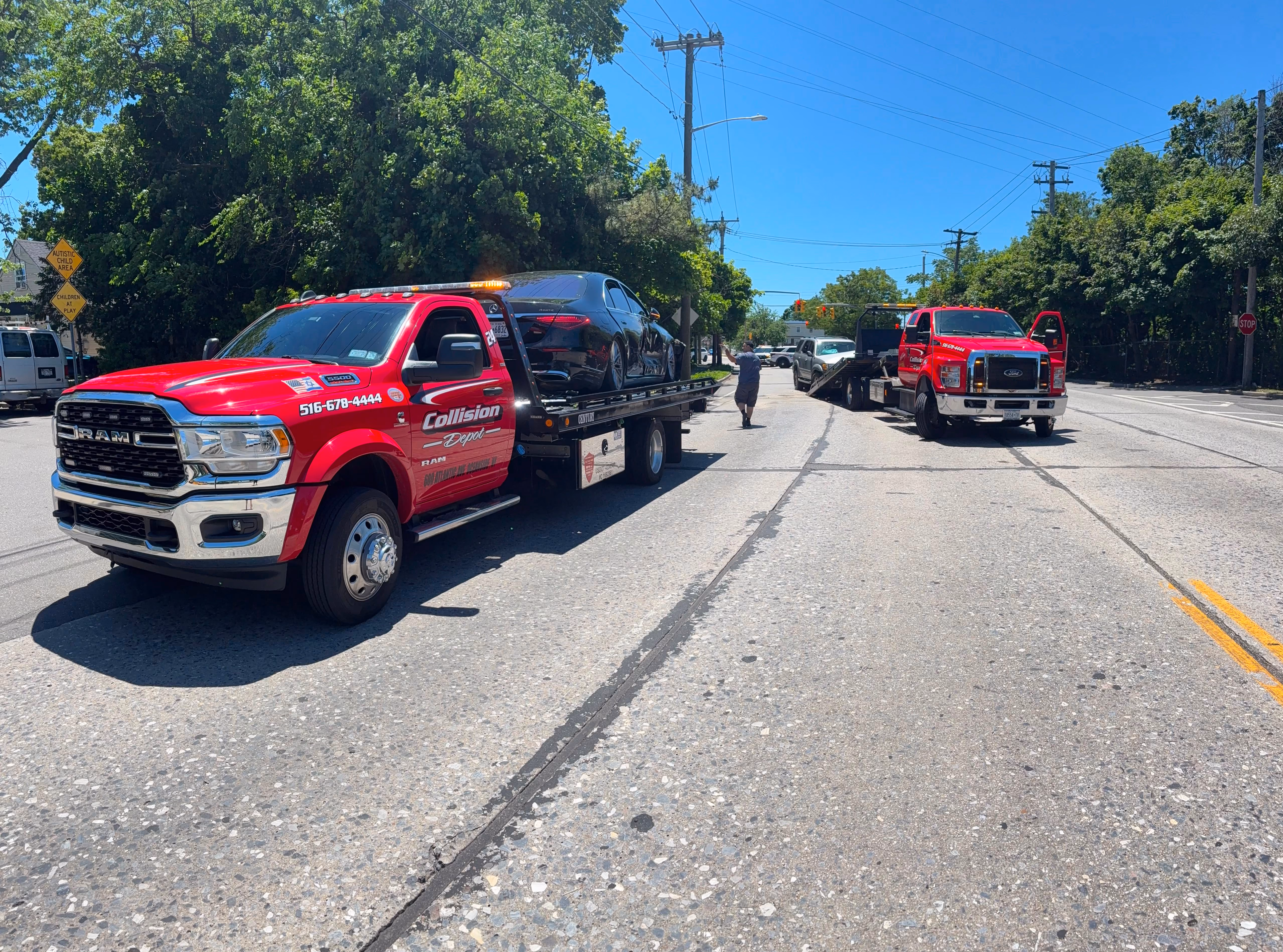Two red tow trucks on a sunny street, one carrying a black car, a person walking between the trucks, with green trees and road signs in the background.