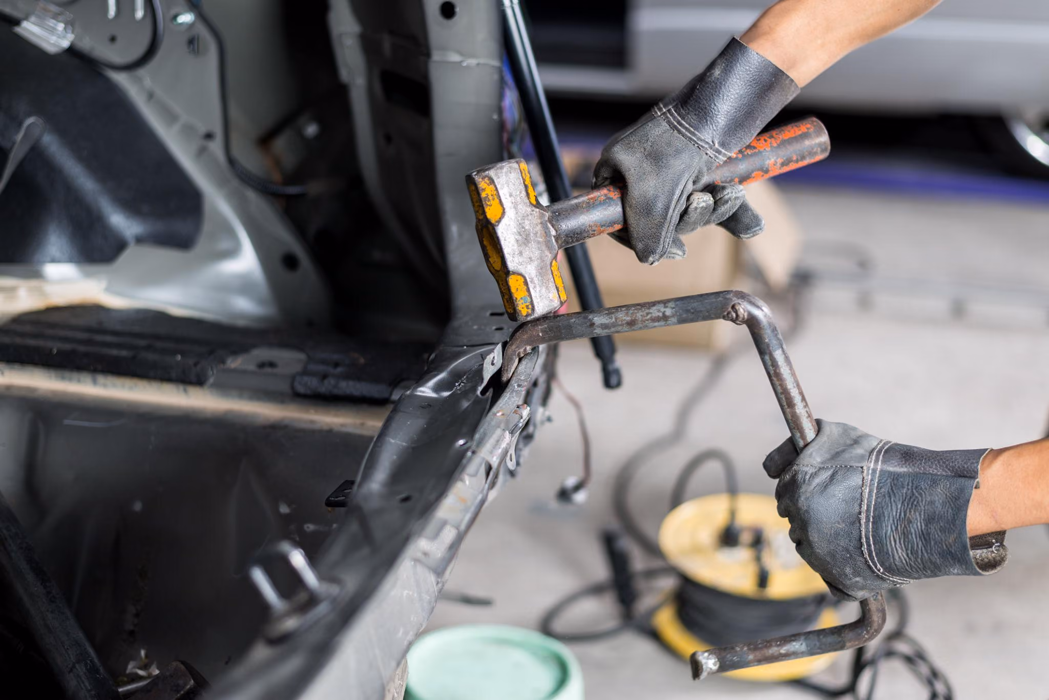 Person wearing black gloves using a hammer and a metal tool to repair or bend a damaged section of a car frame.