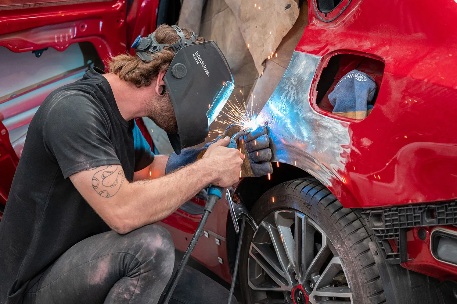 Man wearing a black welding helmet and gloves kneels while welding a metal panel on the rear side of a red car with sparks flying from the welding area.