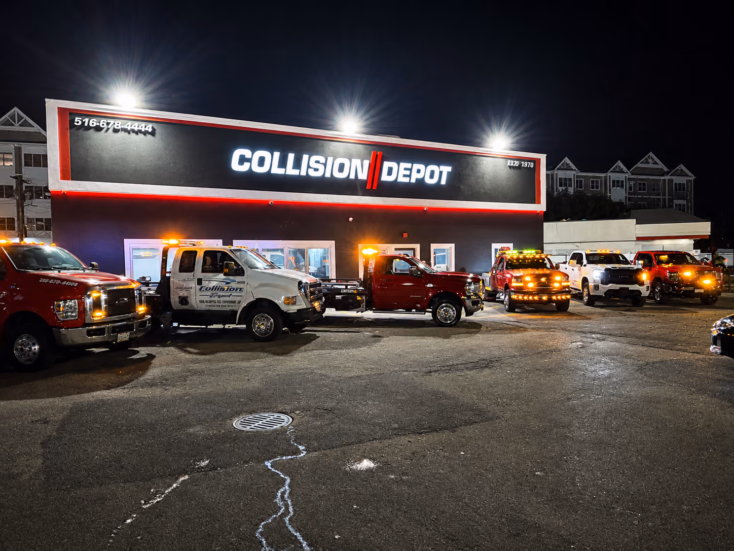 Night view of Collision Depot building with illuminated sign and six tow trucks and service vehicles with flashing lights parked in front on asphalt.