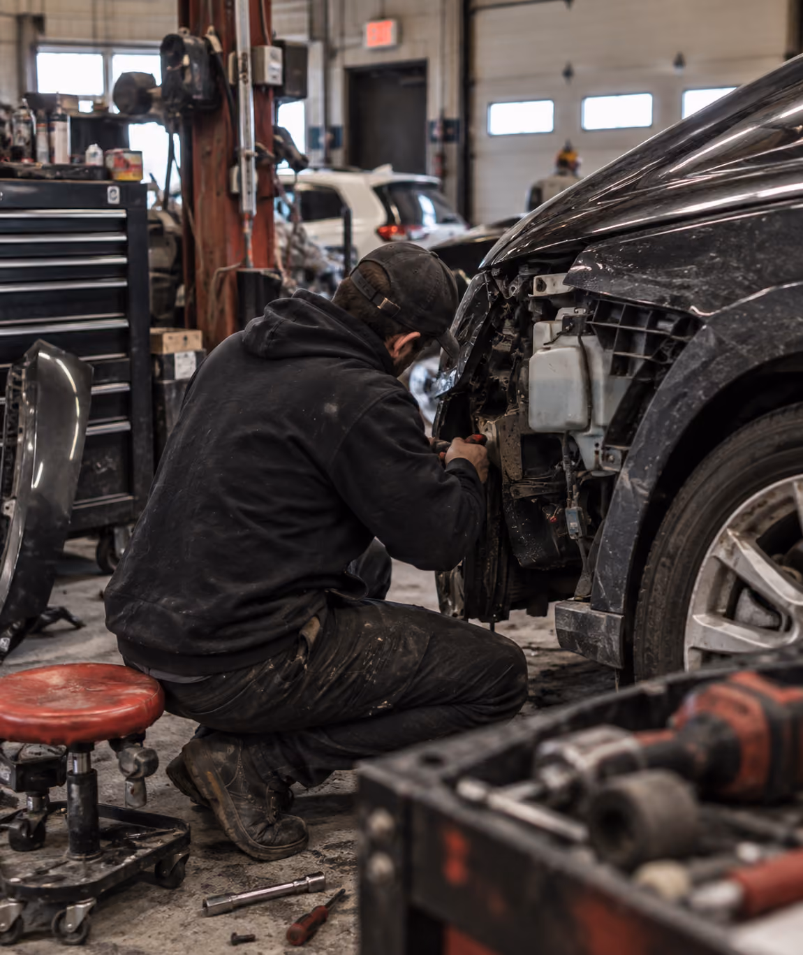 A mechanic wearing a black cap and dark clothing is kneeling and working on the damaged front end of a black car inside a busy auto repair shop with tools and car parts visible around.