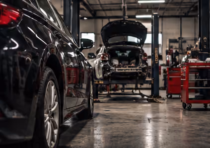 Black car parked inside an auto repair shop with another car raised on a lift being repaired in the background.