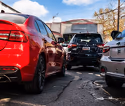 Close-up view of a red car parked on a cracked asphalt surface with a black car and a white car nearby, under a partly cloudy sky.