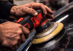 Close-up of hands using a red and black electric buffer machine to polish a black car surface.