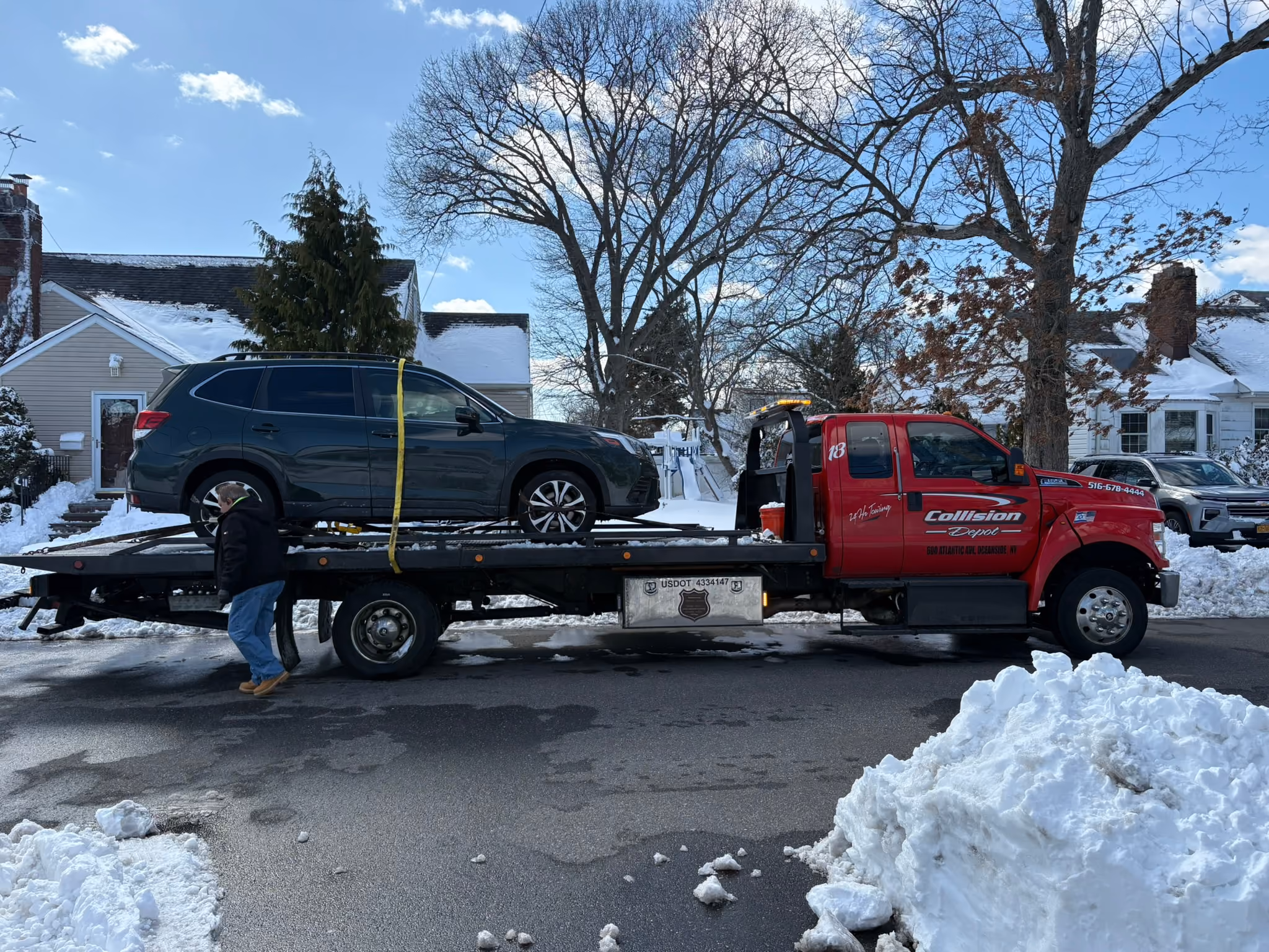 A red Collision Depot tow truck parked on a snowy street with a dark green SUV loaded on its flatbed, a man walking near the truck wearing a black jacket and blue jeans, houses and trees in the background with patches of snow on rooftops and ground.