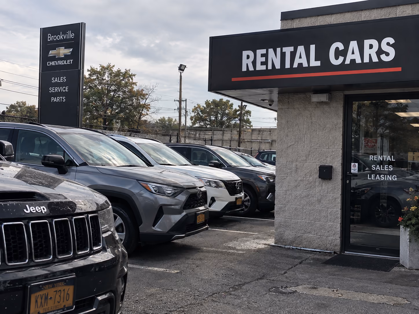 Parking lot of a car rental business with a sign reading 'RENTAL CARS' above the entrance. Several parked cars including a Jeep and other SUVs are visible. A tall sign for Brookville Chevrolet indicates sales, service, and parts. The scene is outdoors under a cloudy sky, with trees and utility poles in the background.