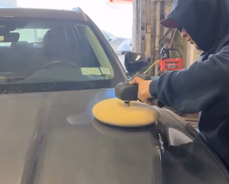 Person wearing a hoodie and cap using a polishing machine with a yellow pad on the hood of a black car inside a garage.