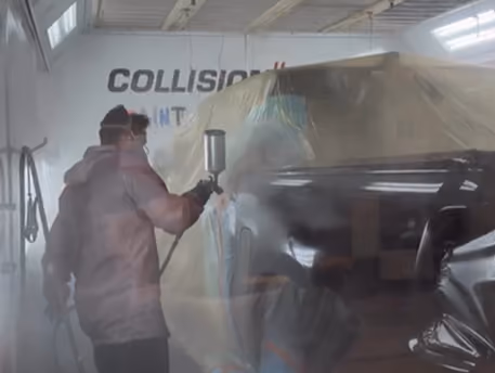 Person wearing protective gear spray painting a vehicle in a workshop with the word 'COLLISION' partially visible on the wall behind.