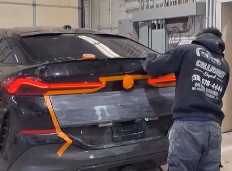 Person working on the rear of a black SUV inside a garage, preparing for vehicle repair; taillights are on and the rear license plate area is masked with gray material and orange tape.
