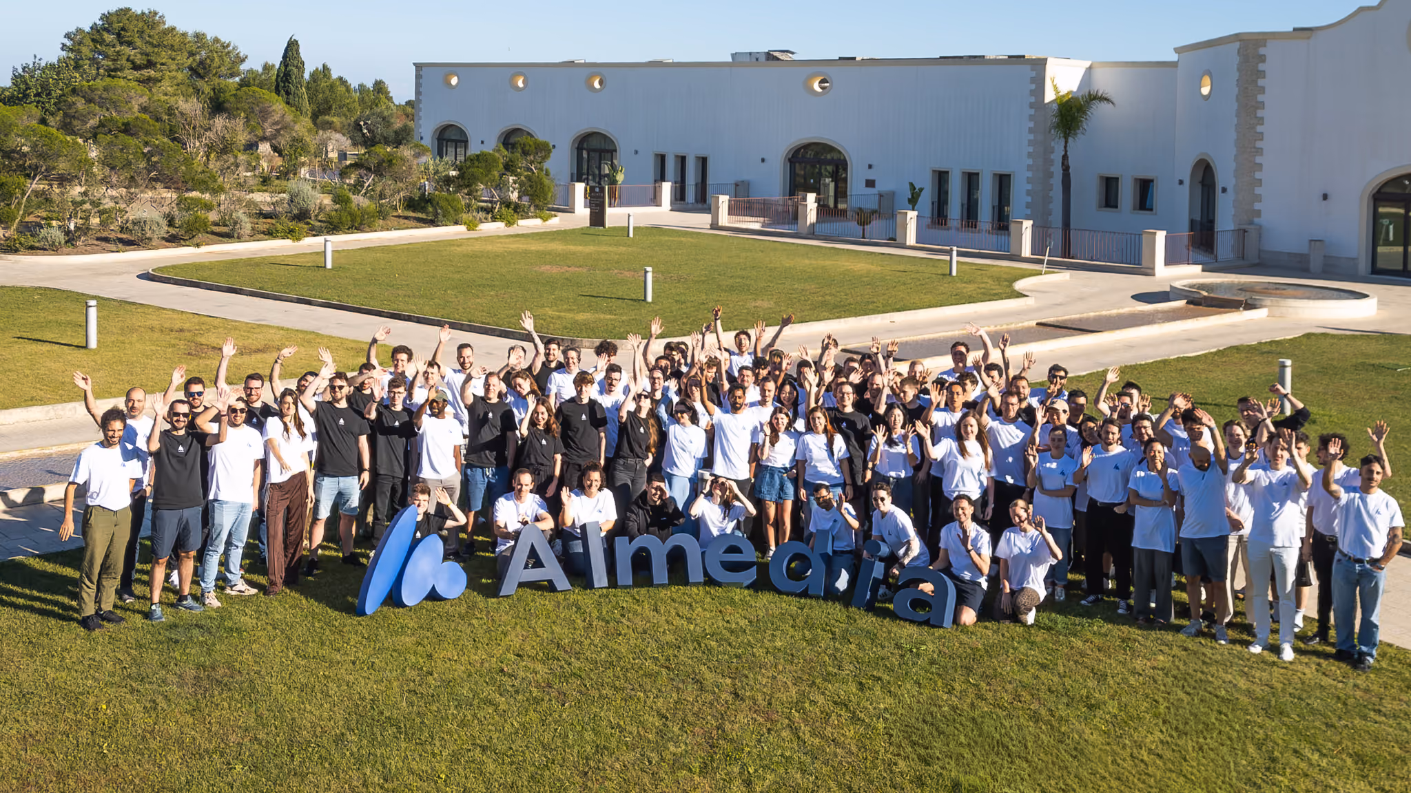 Full team of Almedia standing on a lawn waving at the camera, with a big Almedia logo in front of them