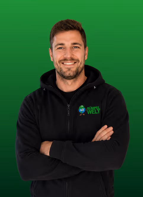 Smiling man with beard wearing black jacket with Rümpel Welt logo on green background.