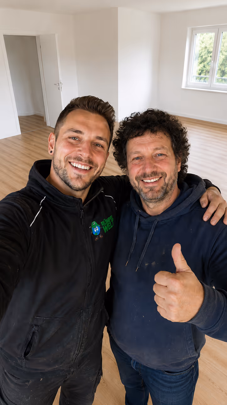 Two smiling men in work clothes posing for selfie in an empty room, one showing a thumbs-up.