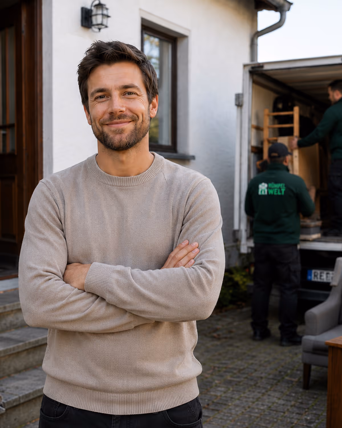 Smiling man in beige sweater with arms crossed in front of house while movers load a truck in background.