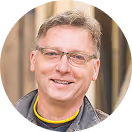 Smiling man with glasses wearing a dark jacket and yellow shirt against a blurred background.