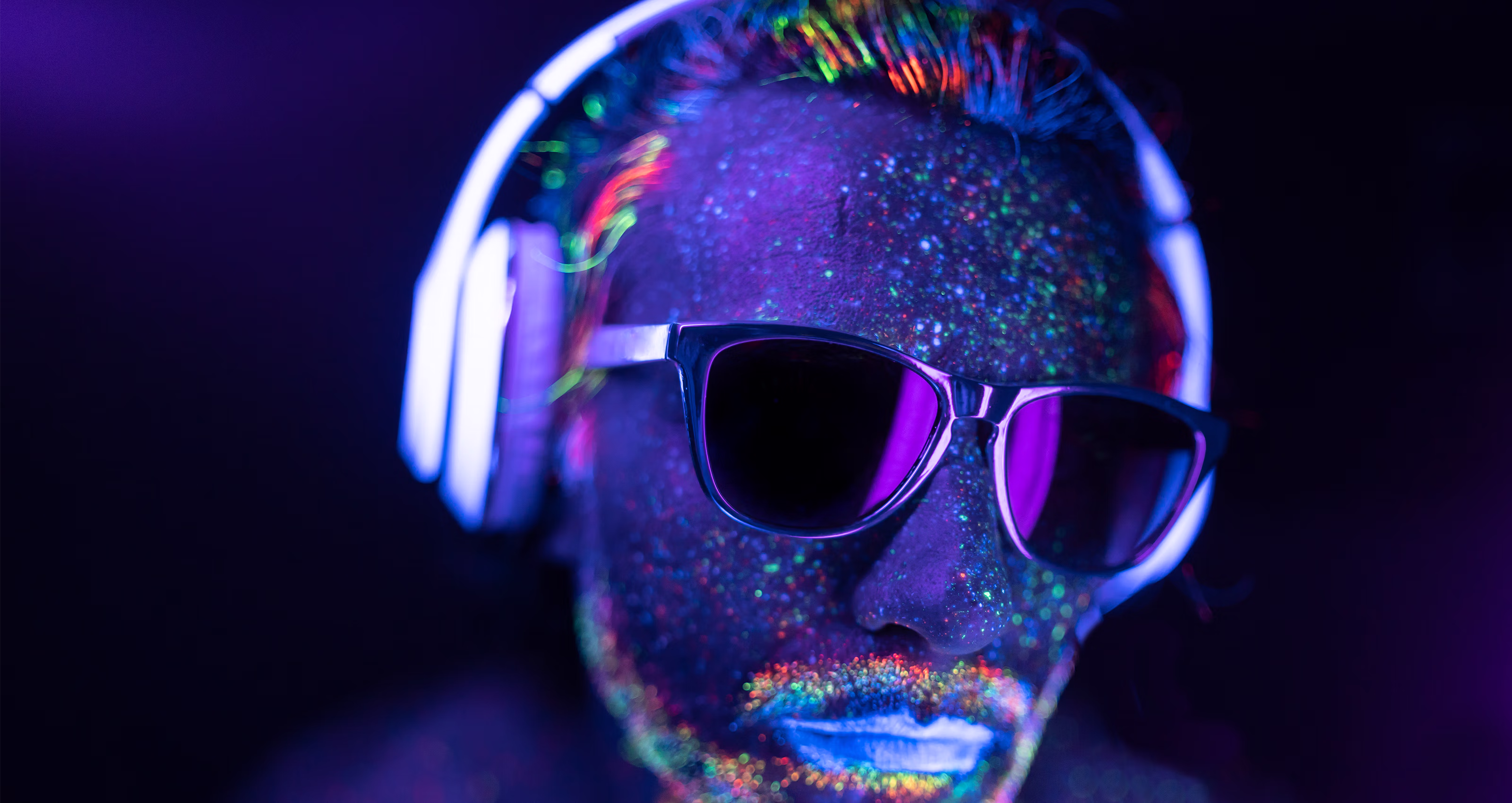 Person with neon glowing paint on face wearing sunglasses and white headphones against a dark background.