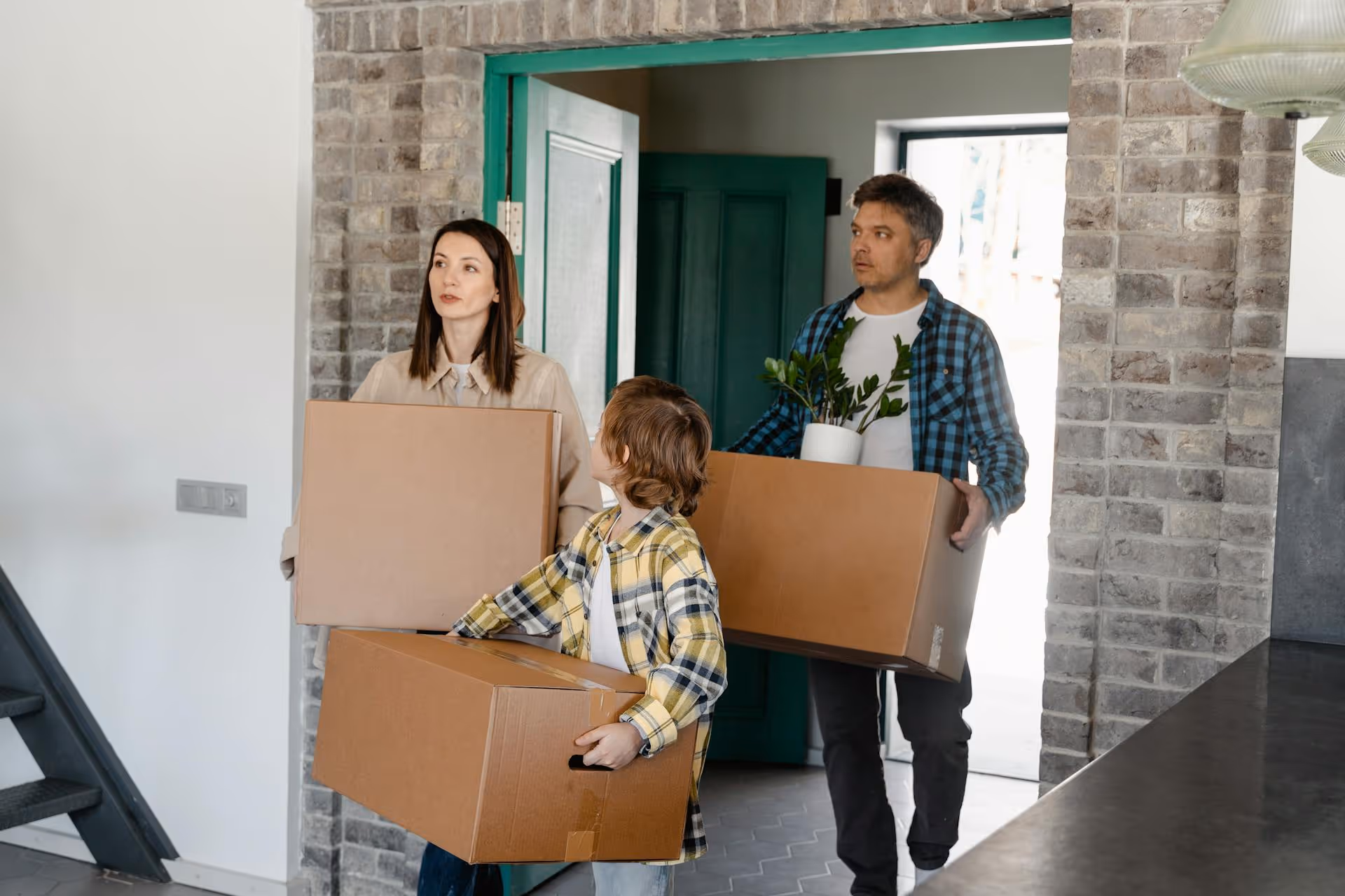 Family of three carrying cardboard boxes while entering a home through a green door.
