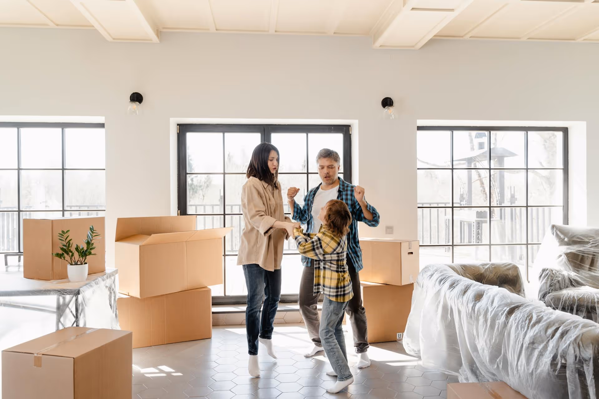 Family of three dancing together in a sunlit room surrounded by moving boxes and furniture covered in plastic.