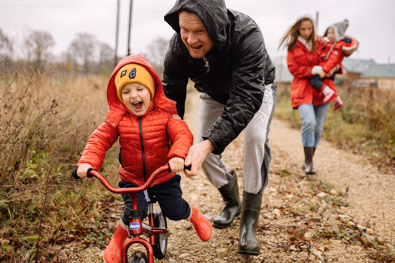 Smiling father in a black jacket helping his excited young child ride a red bike on a gravel path while a woman in a red coat carries another child in the background.