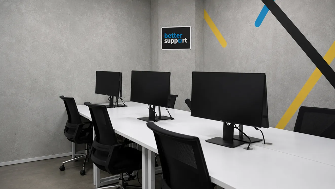 Modern office workspace with white desks, black chairs, monitors, and a wall sign reading 'better support'.