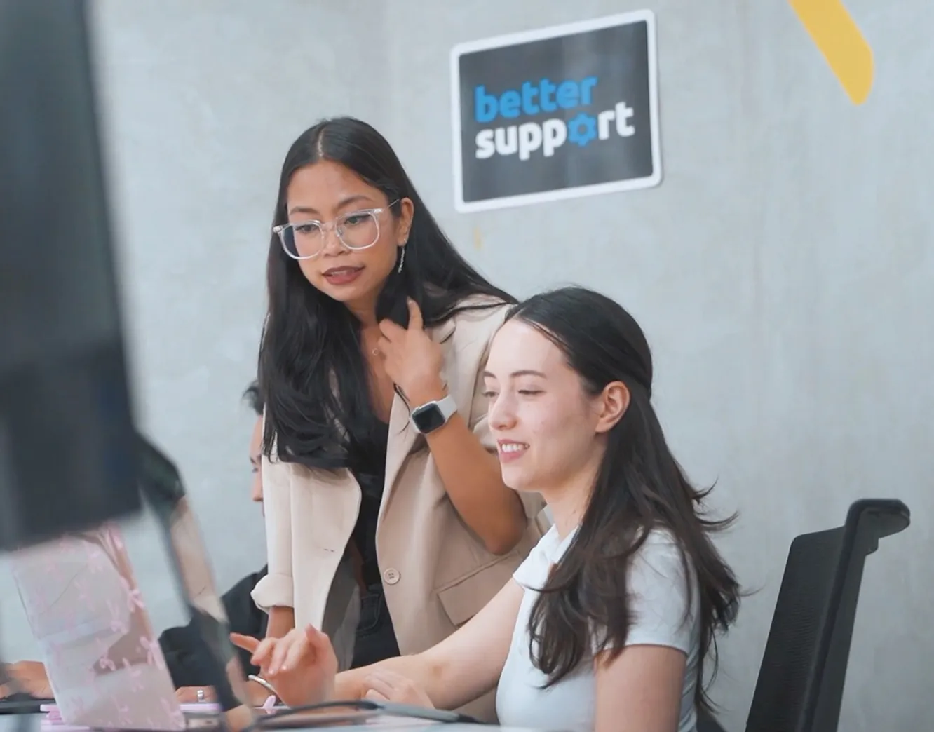 Two women working together at a computer with a sign reading 'better support' on the wall.