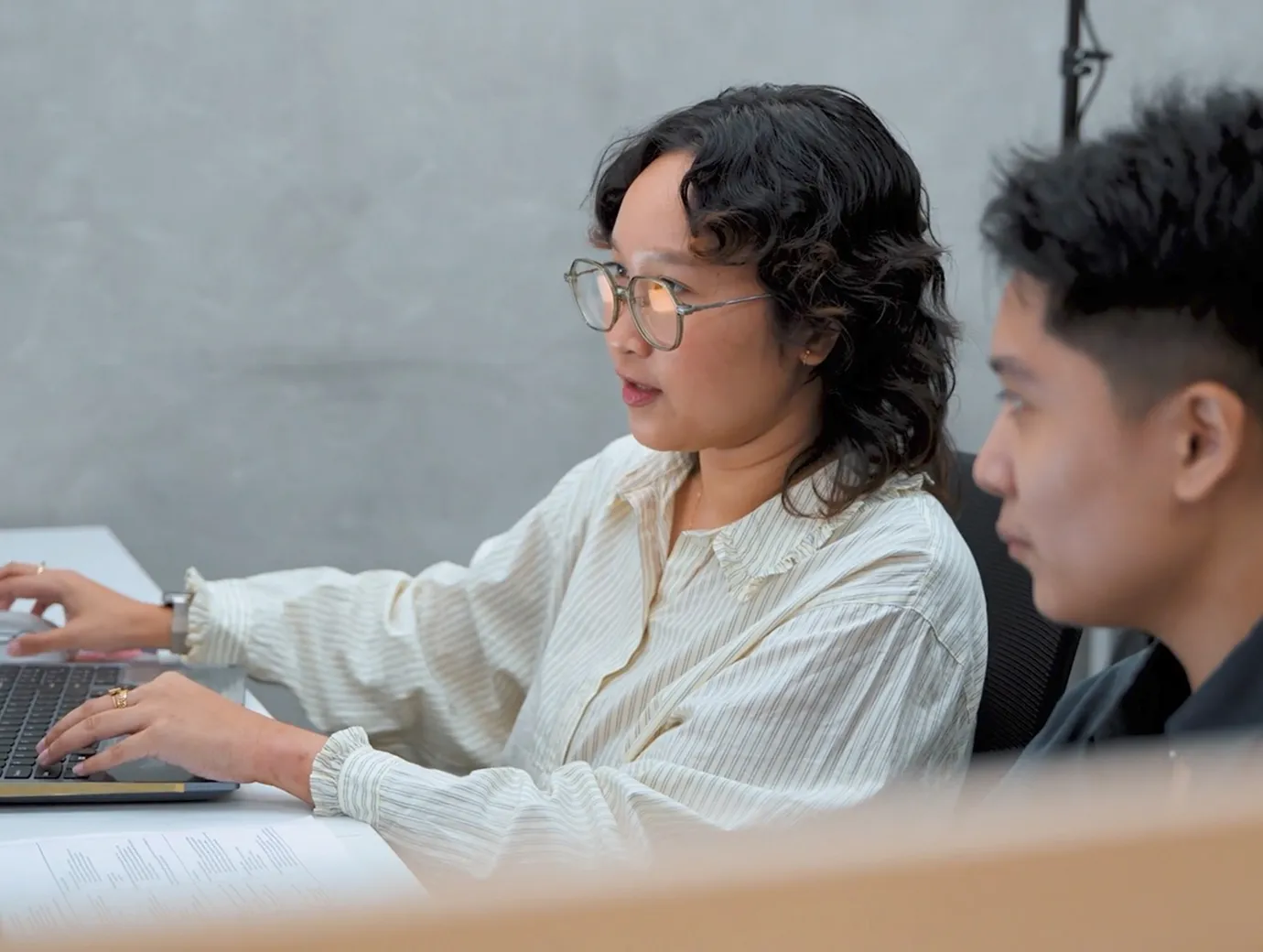 Two people in discussion, one using a laptop keyboard while sitting at a desk with papers.