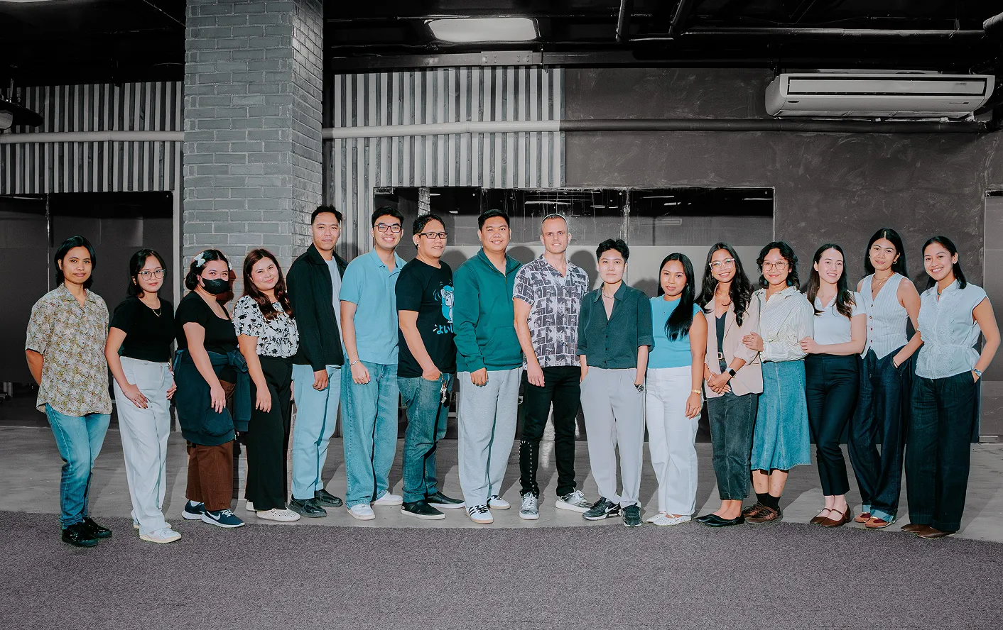 A group of 15 adults standing indoors in a line posing for a photo against a gray brick and wall background.