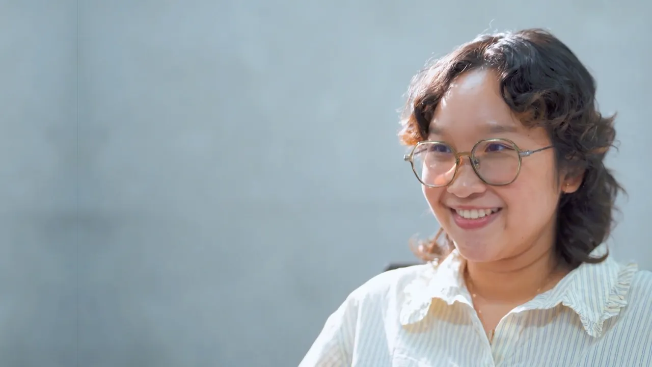 Smiling woman with glasses and short curly hair wearing a striped shirt against a gray background.