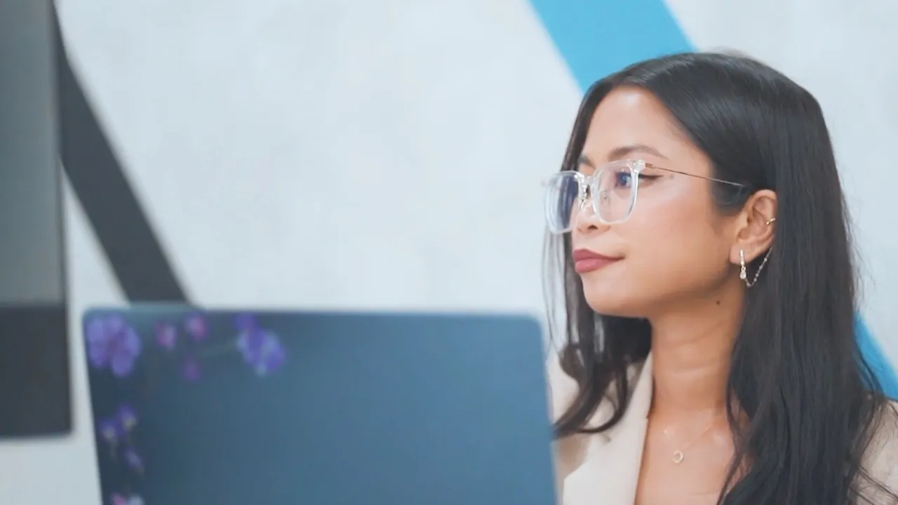 Woman wearing clear glasses working on a laptop with a blue and gray background.