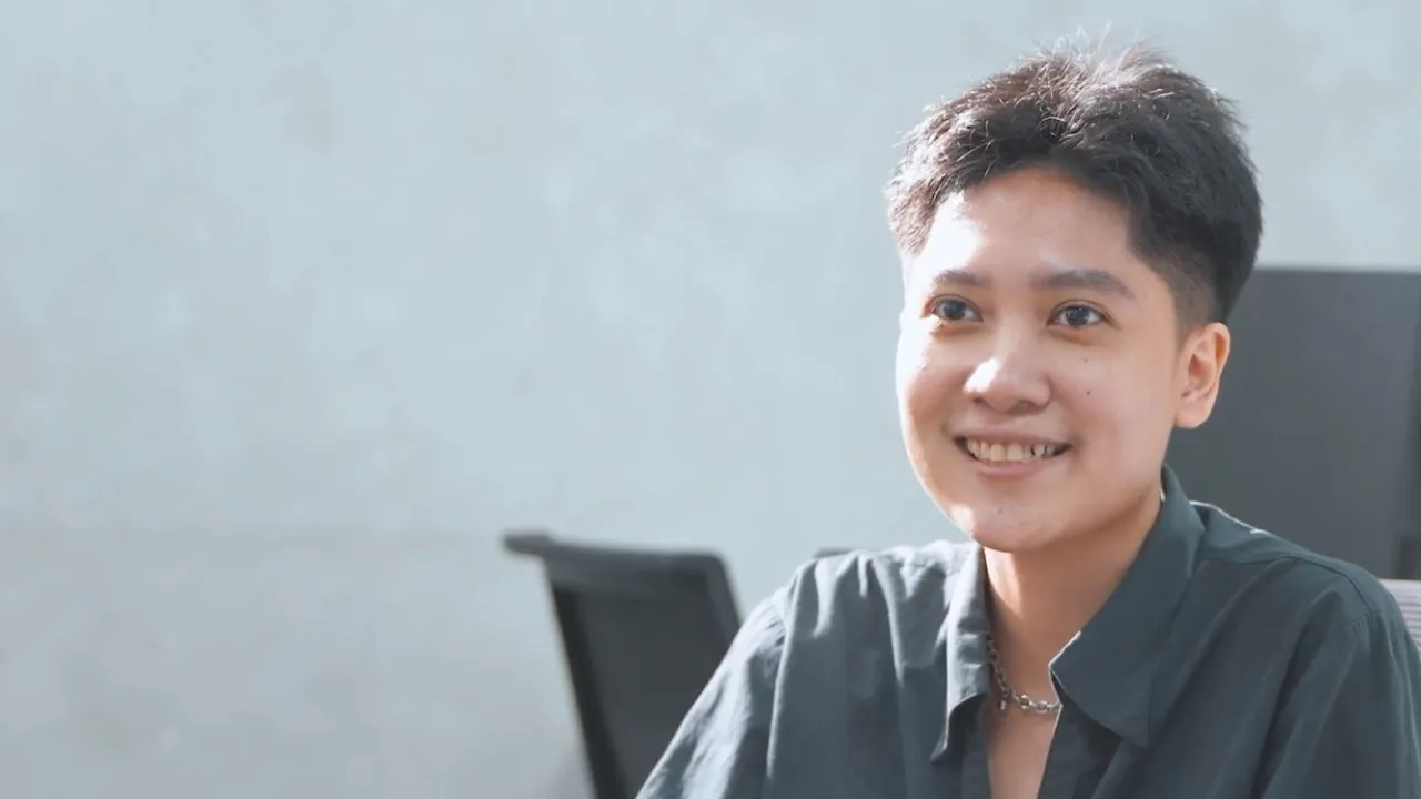 Young person with short hair smiling, wearing a dark shirt and silver chain necklace indoors.