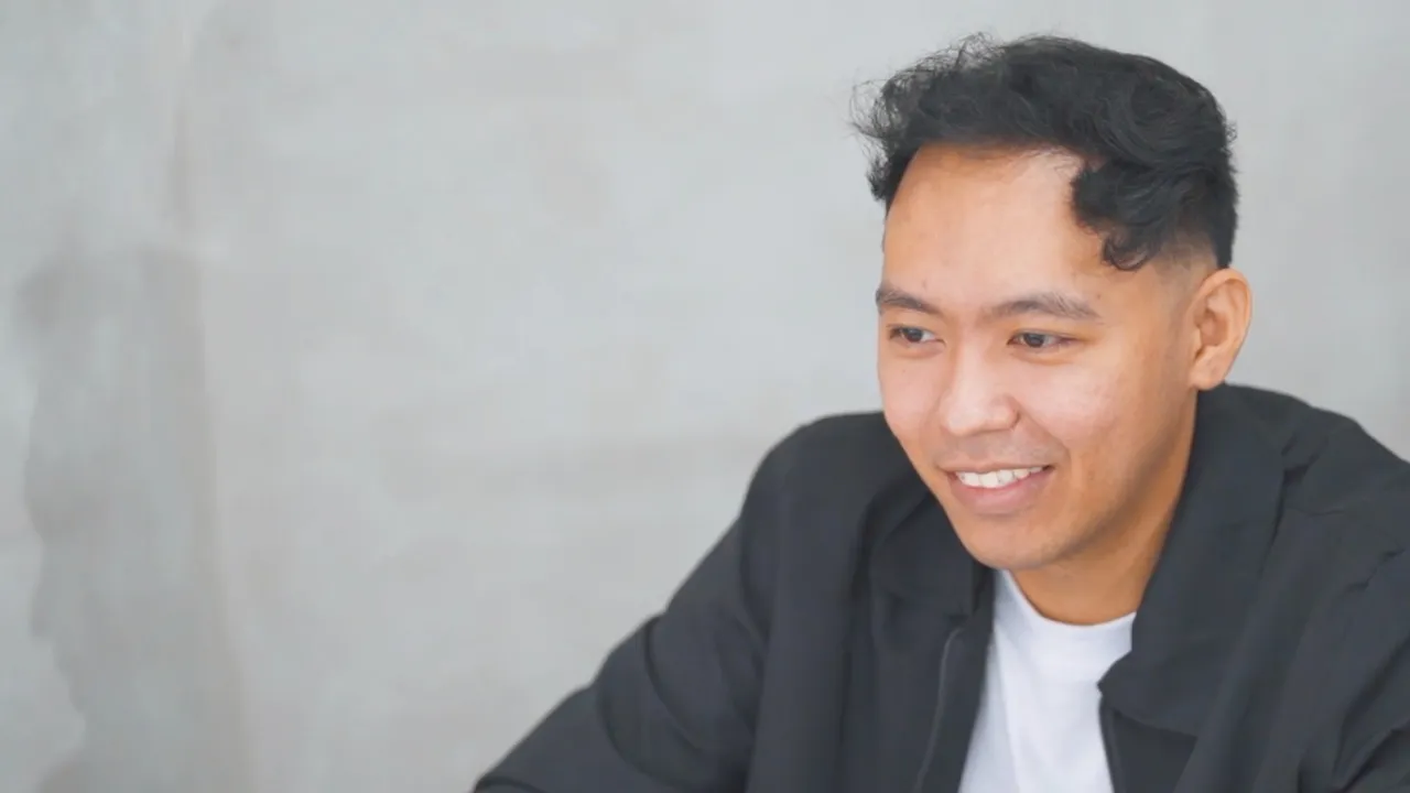Smiling young man with short black hair wearing a black jacket and white shirt against gray wall.