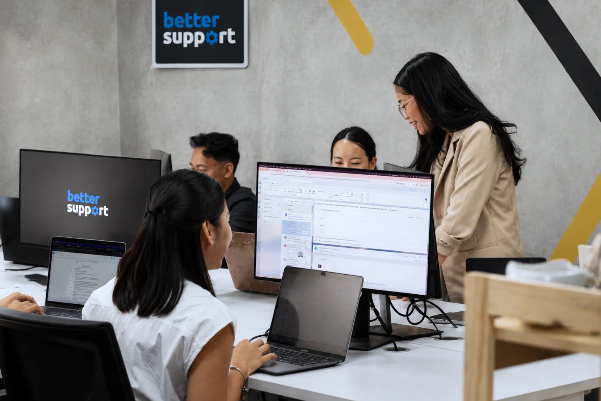 Four coworkers collaborate around desks with laptops and monitors in an office with a ‘better support’ sign.