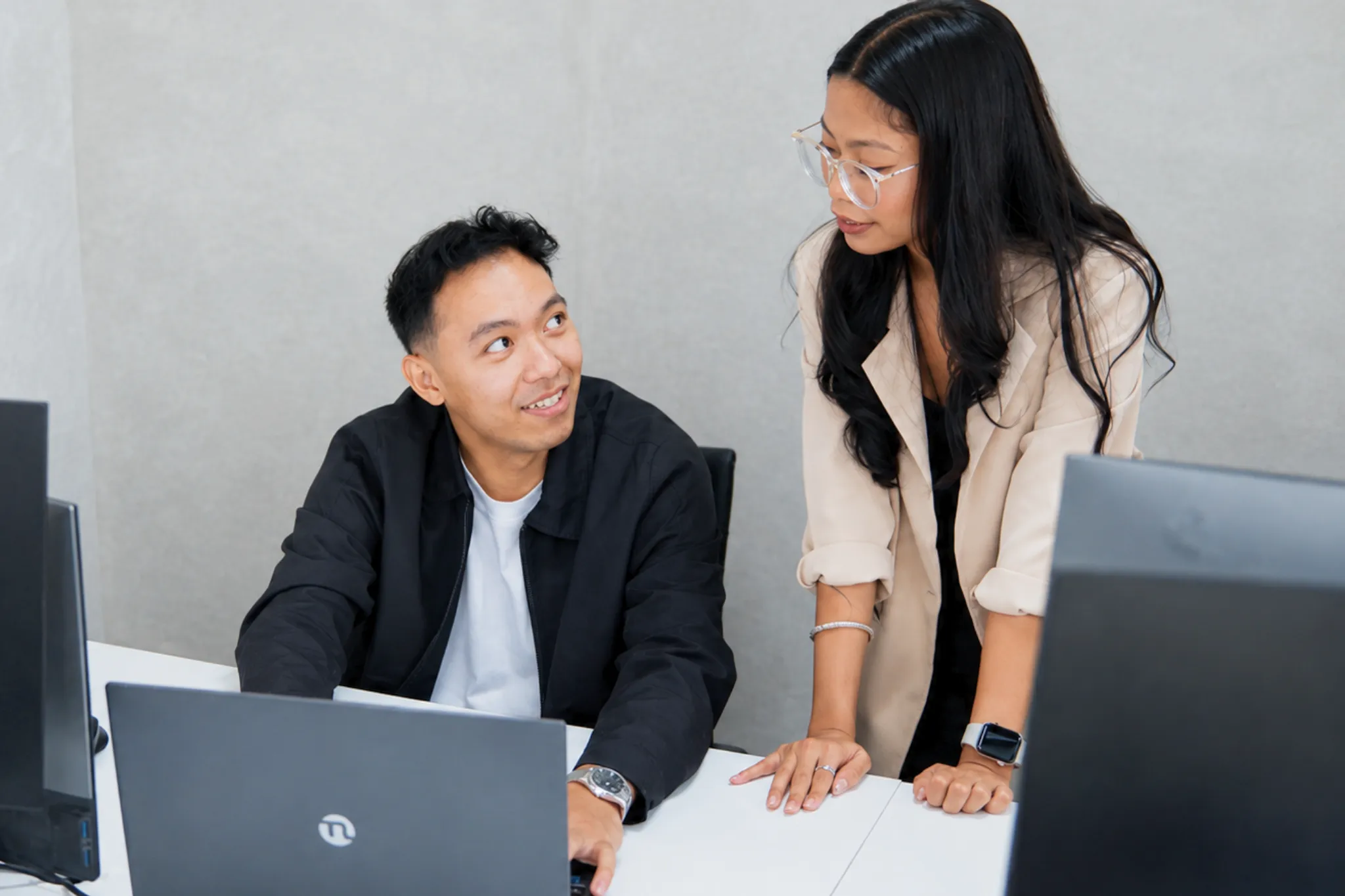 Young man sits at desk with laptop, woman stands beside him talking, both indoors at work.