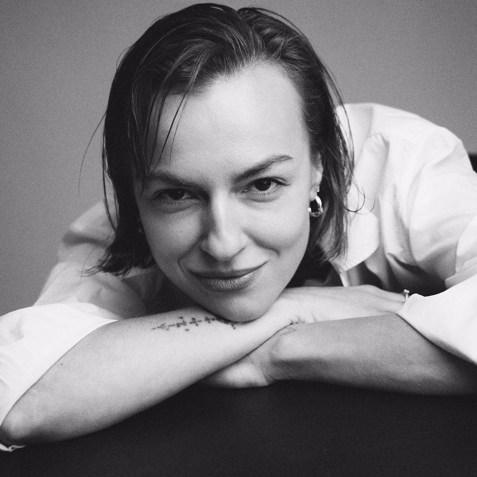 Black and white close-up of a smiling woman resting her head on folded arms with a tattoo on her forearm.
