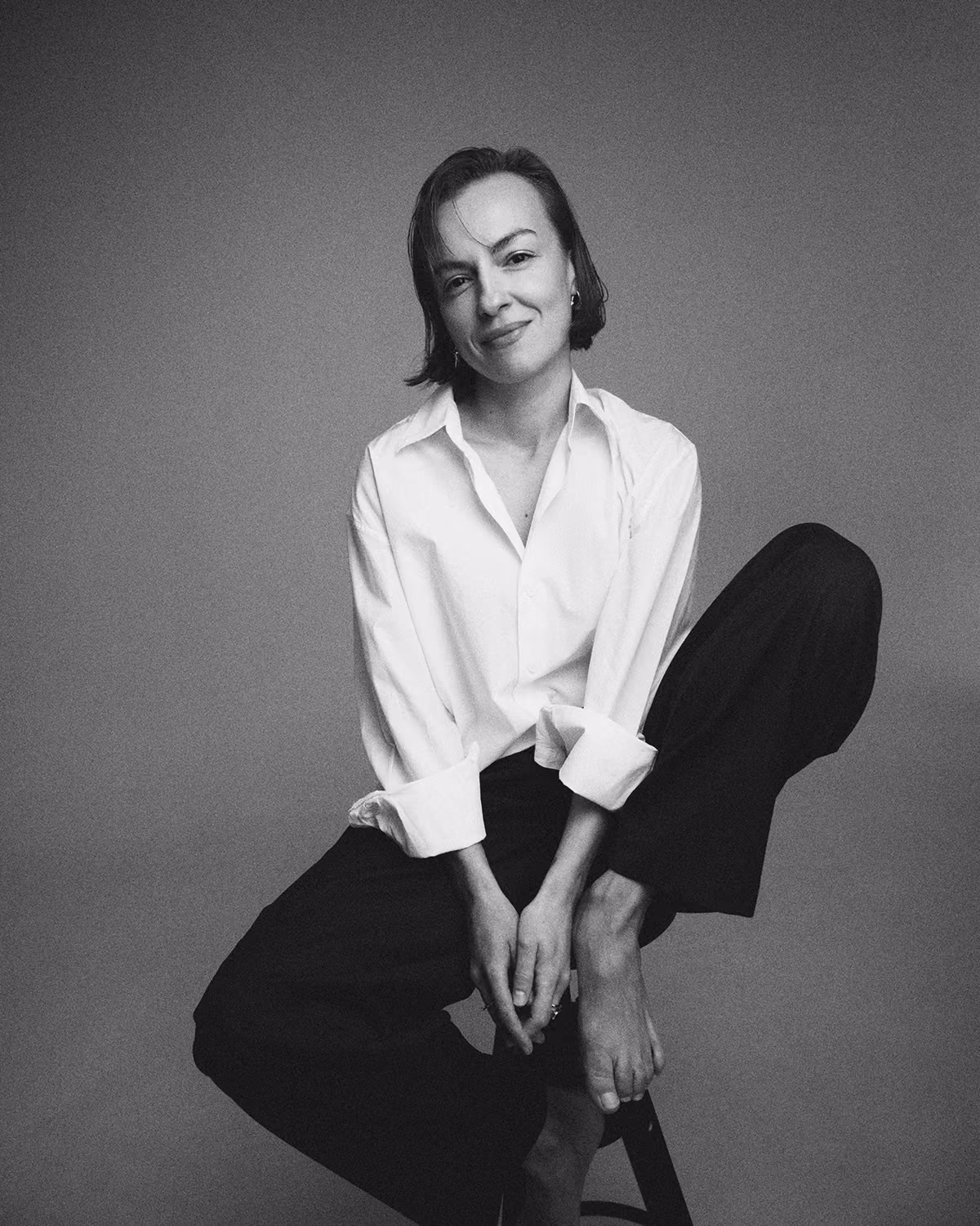 Smiling woman with short hair wearing a white shirt and black pants sitting barefoot on a stool.