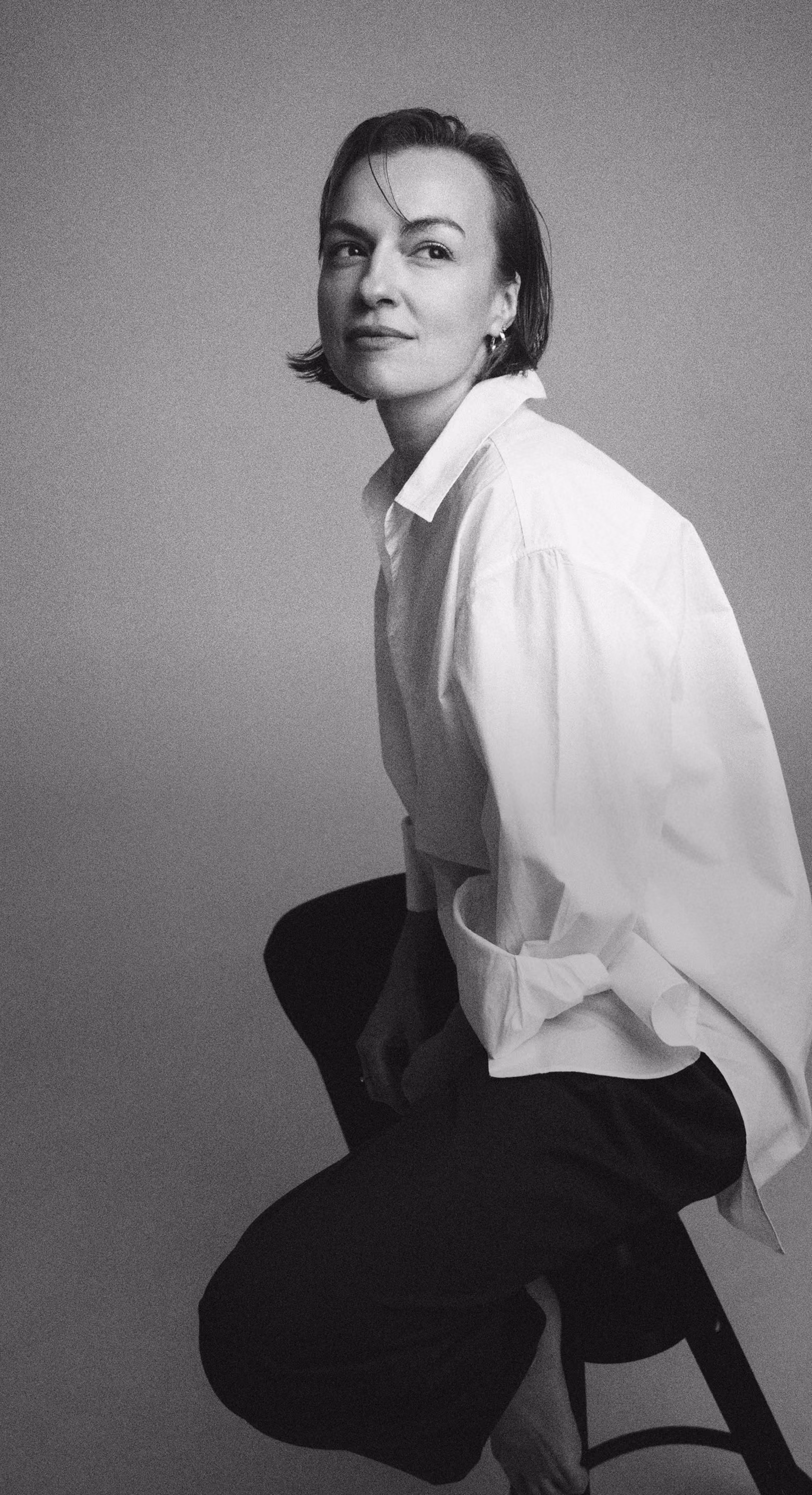 Black and white portrait of a woman sitting on a stool, wearing a white shirt and dark pants.
