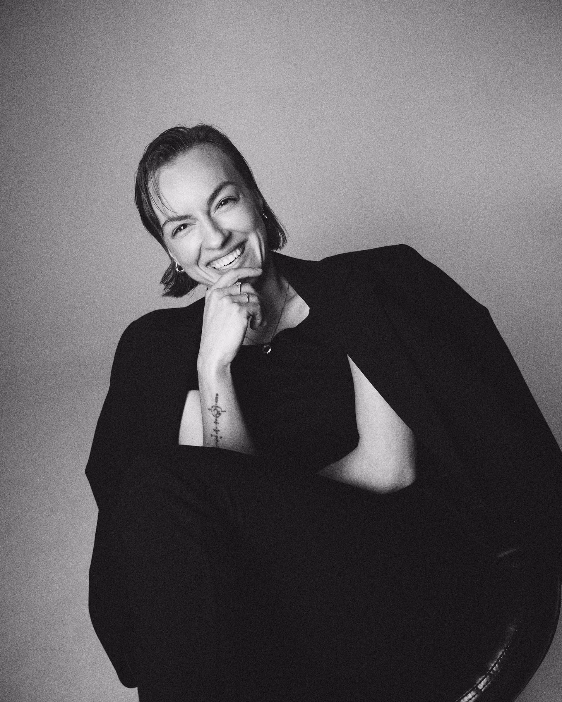 Black and white portrait of a smiling person with short hair, resting chin on hand, wearing dark clothes.