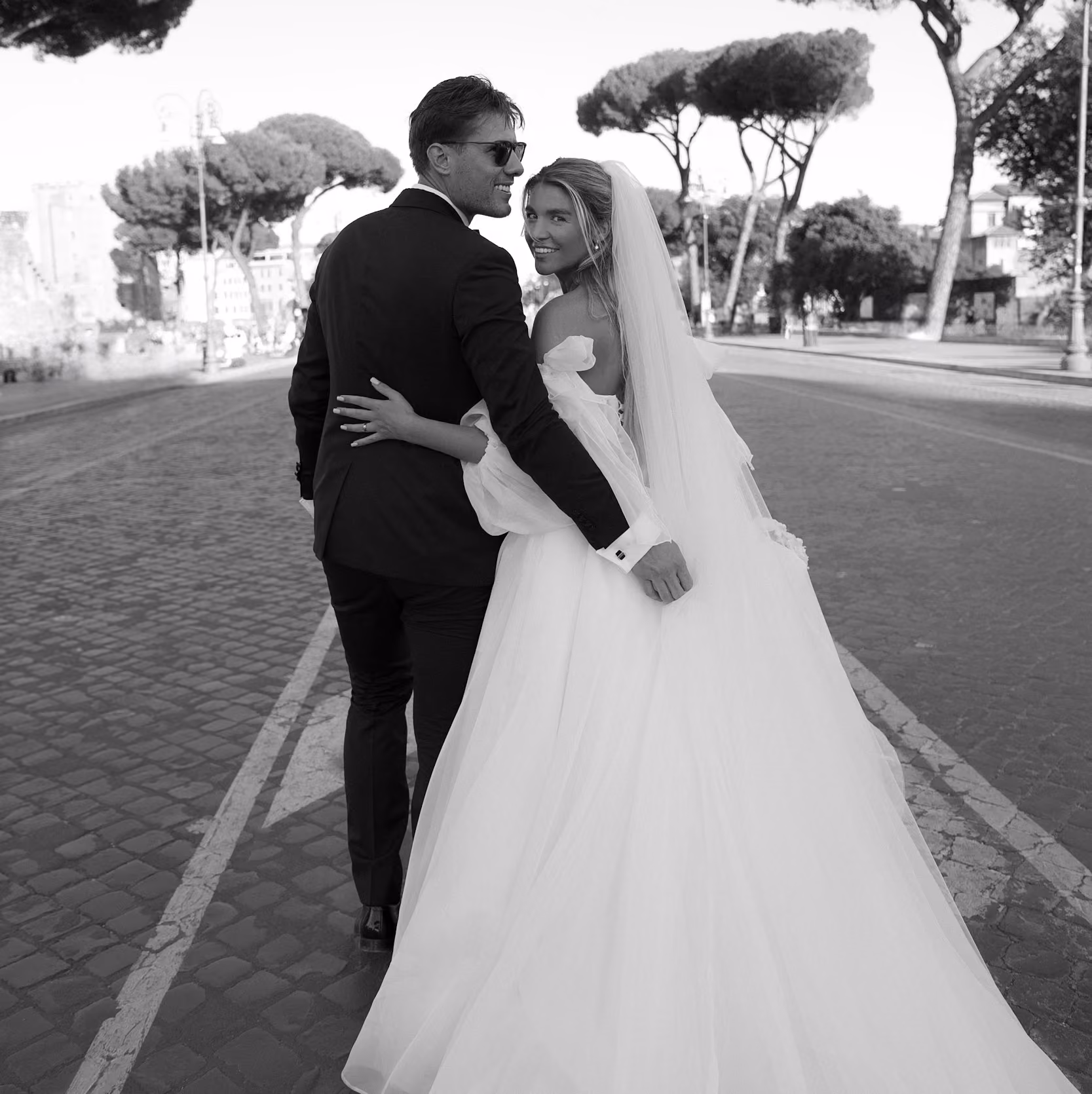 Bride and groom walking arm in arm on cobblestone street, both smiling and looking back.
